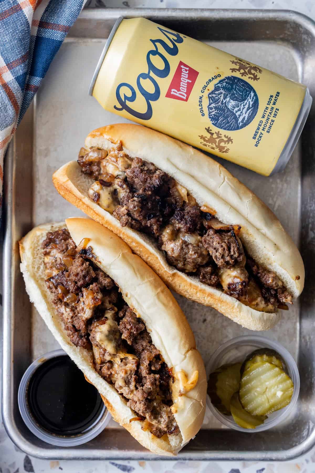 Two cheesesteak sandwiches with ground beef and melted cheese on a tray resemble the classic Blackstone chopped cheese, served with pickles, a cup of sauce, and a can of Coors Banquet beer.