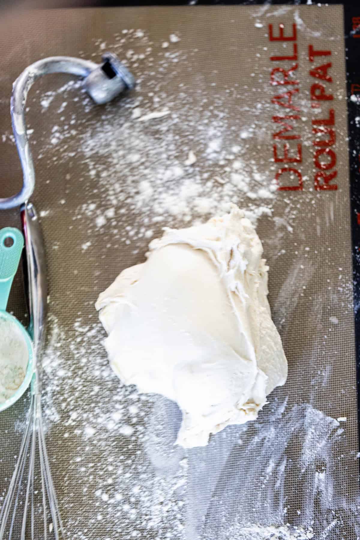 A mound of dough on a floured silicone baking mat awaits shaping into classic Scottish Morning Rolls, with a measuring cup, whisk, and dough hook nearby.