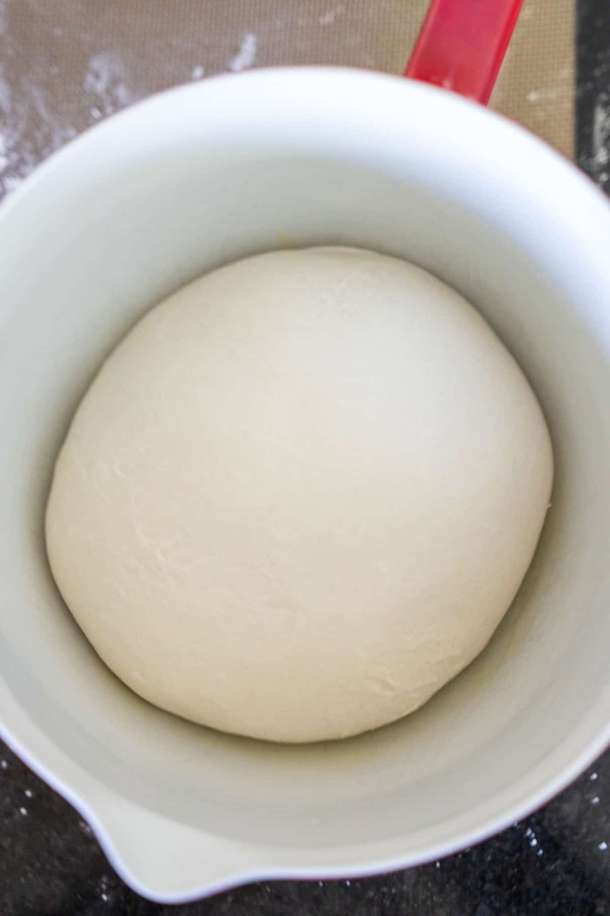 A ball of risen dough for Scottish Morning Rolls rests in a white mixing bowl with a red handle, placed on a dark countertop.