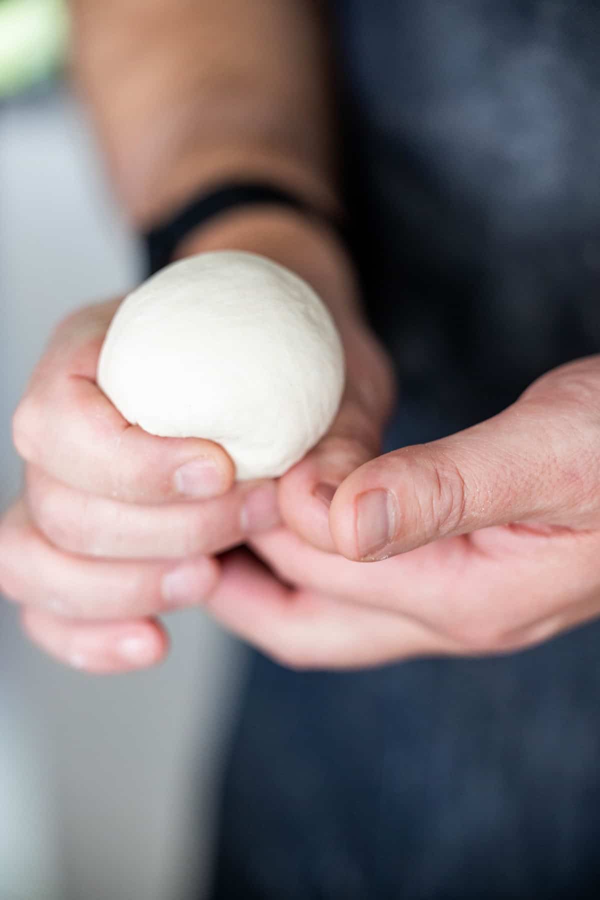 A person holding a smooth ball of dough with both hands, preparing to bake classic Scottish morning rolls.