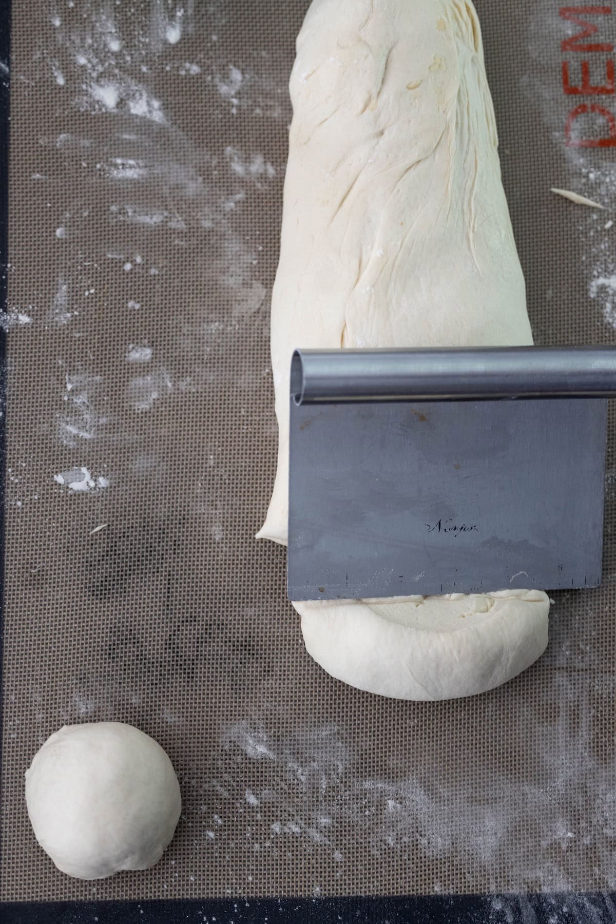 A metal bench scraper cuts a log of dough on a silicone baking mat, preparing portions for Scottish morning rolls; a small ball of dough sits nearby.