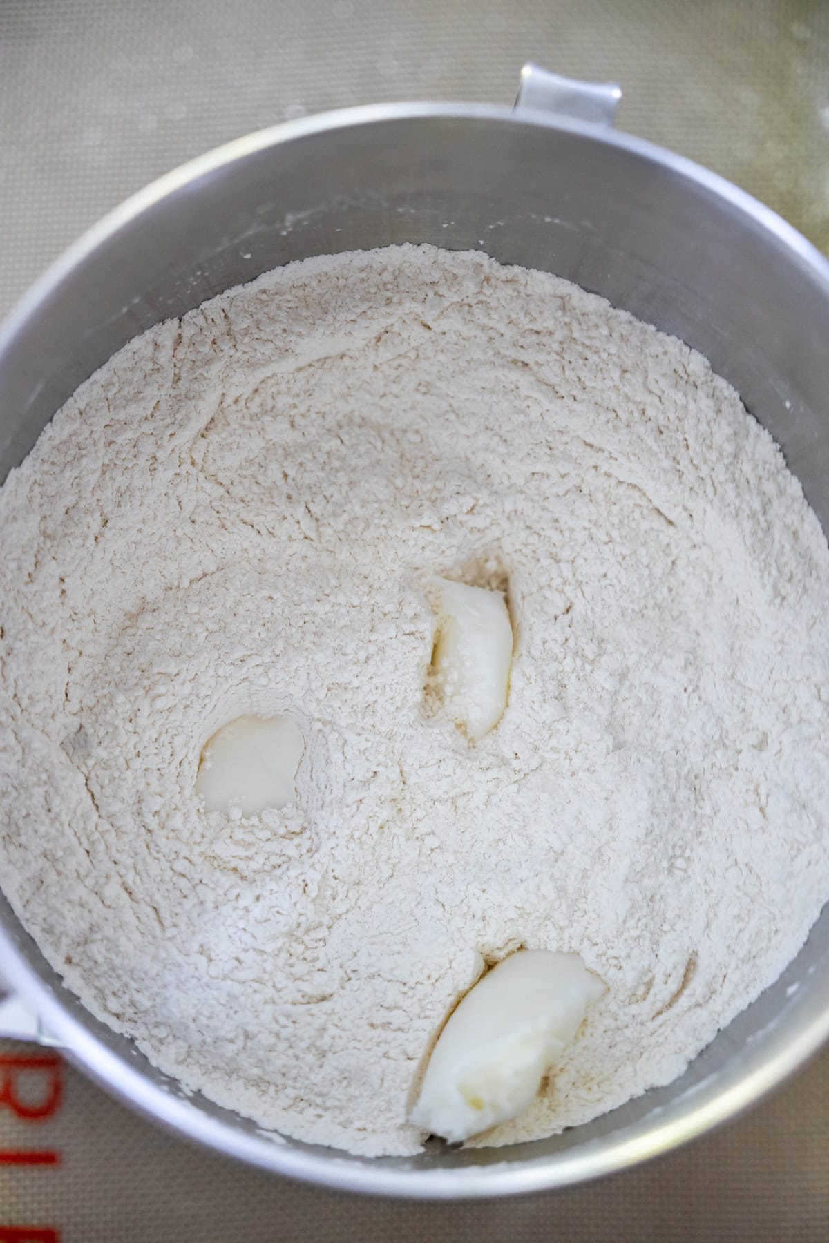 A metal mixing bowl containing flour with three small lumps of solid shortening on top—just the start for baking classic Scottish morning rolls.