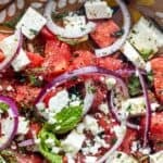 Close-up of a Watermelon Feta Salad with feta cheese cubes, sliced red onions, fresh mint leaves, basil, chopped herbs, and ground black pepper.