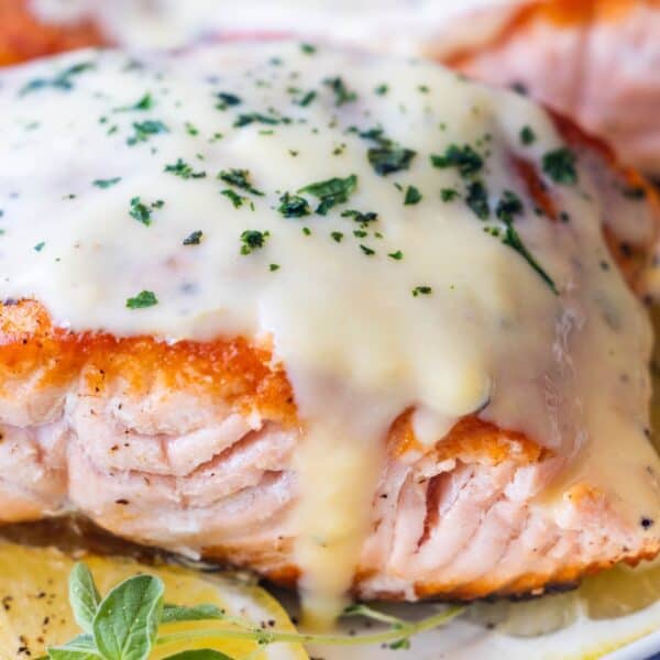 Close-up of Avgolemono Salmon fillet topped with creamy white sauce and garnished with chopped herbs, with a slice of lemon and herb sprig nearby.