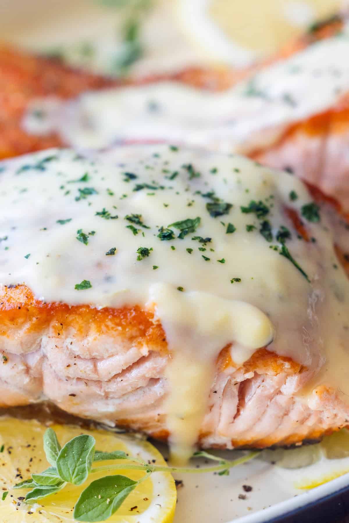 Close-up of cooked Greek salmon fillet topped with creamy white sauce and chopped herbs, garnished with lemon slices and oregano&mdash;an irresistible Greek salmon recipe.