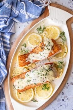 Two pieces of Avgolemono Salmon, topped with creamy sauce and herbs, are served on lemon slices in a fish-shaped dish, with a blue striped towel nearby.