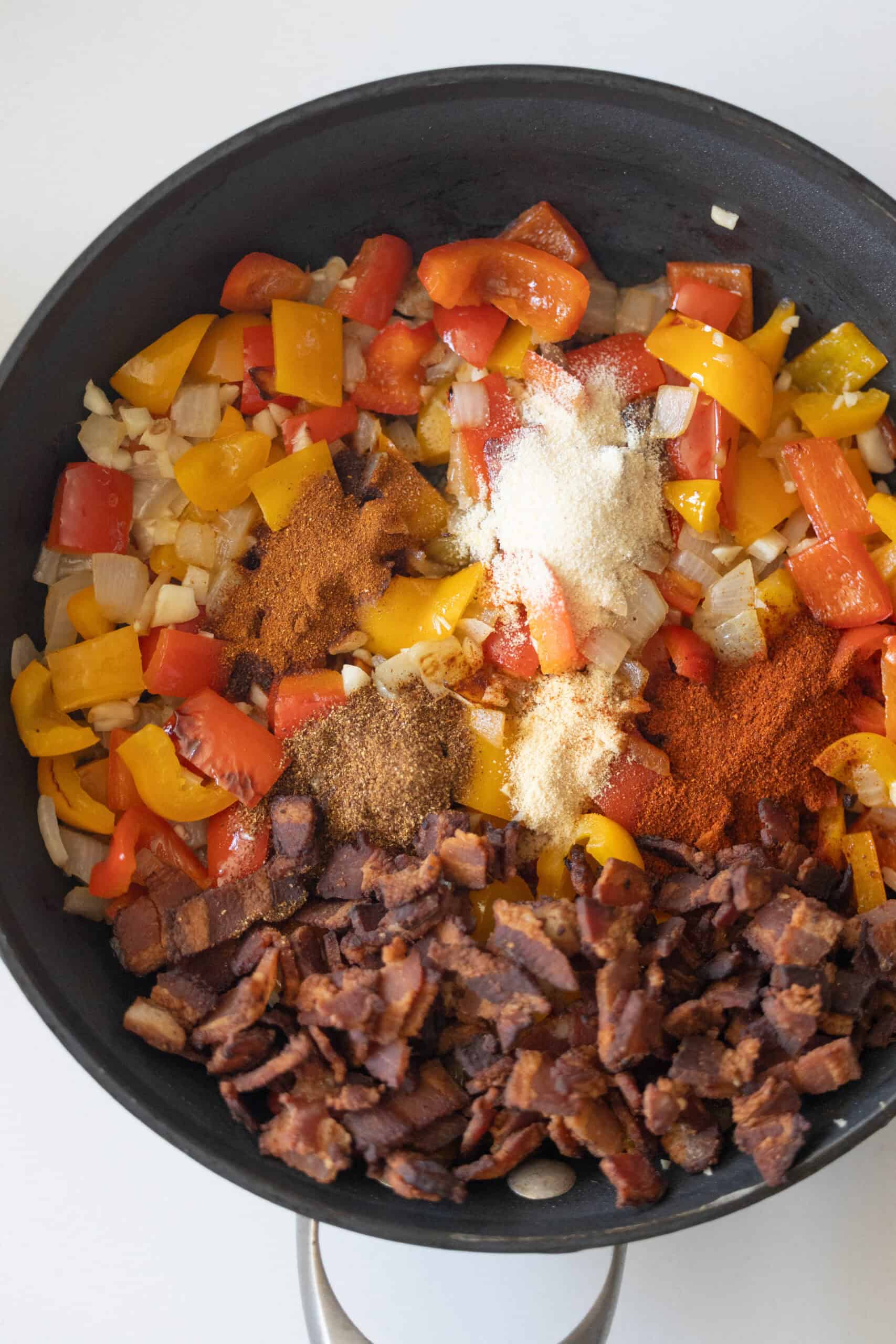 A skillet containing chopped red and yellow bell peppers, onions, crumbled bacon, several piles of assorted spices, and the beginnings of an authentic recipe for flavorful Mexican beans.