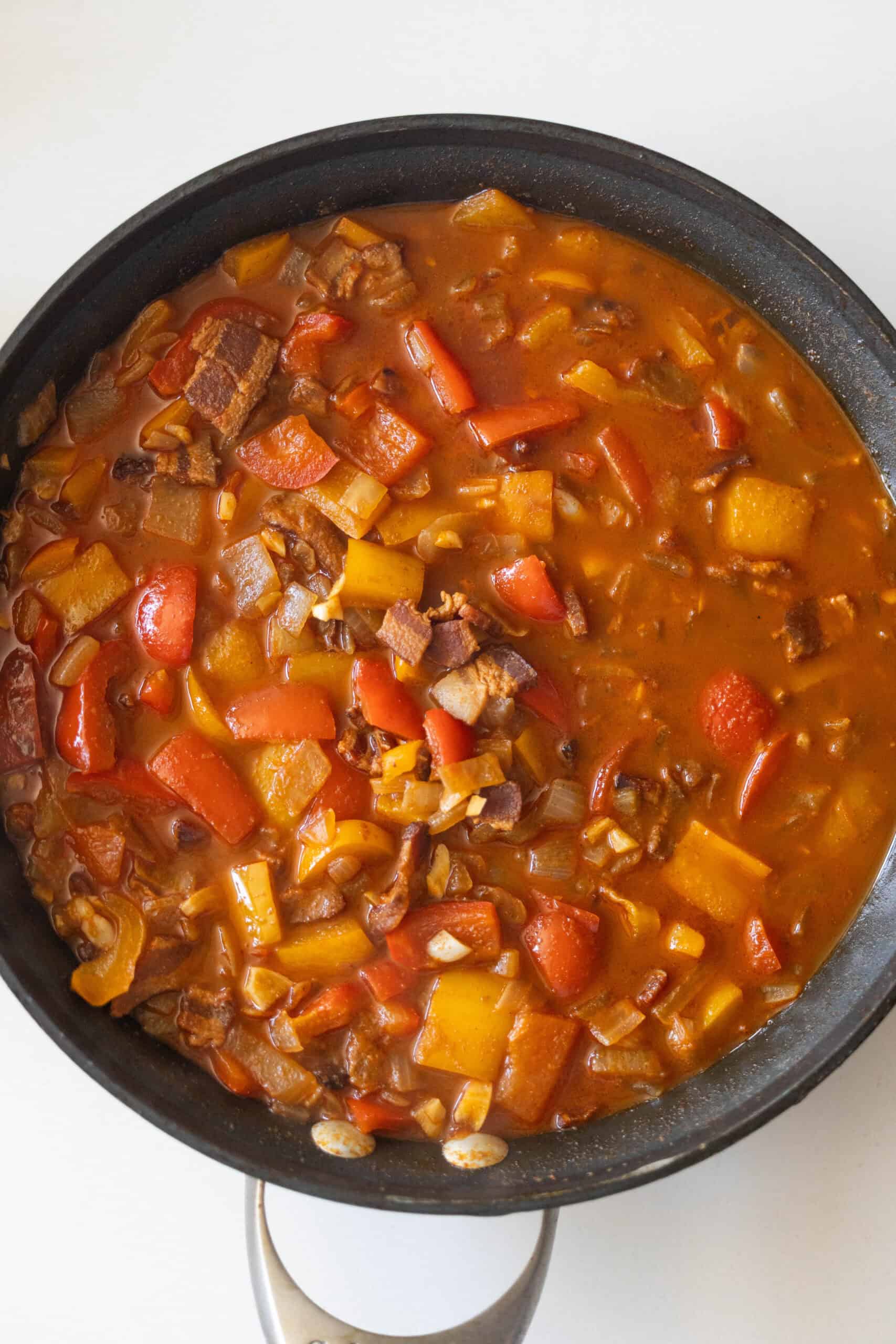 A skillet filled with a tomato-based stew featuring chopped red and yellow bell peppers, bacon pieces, onions, and hearty cowboy beans for a delicious, rustic touch.