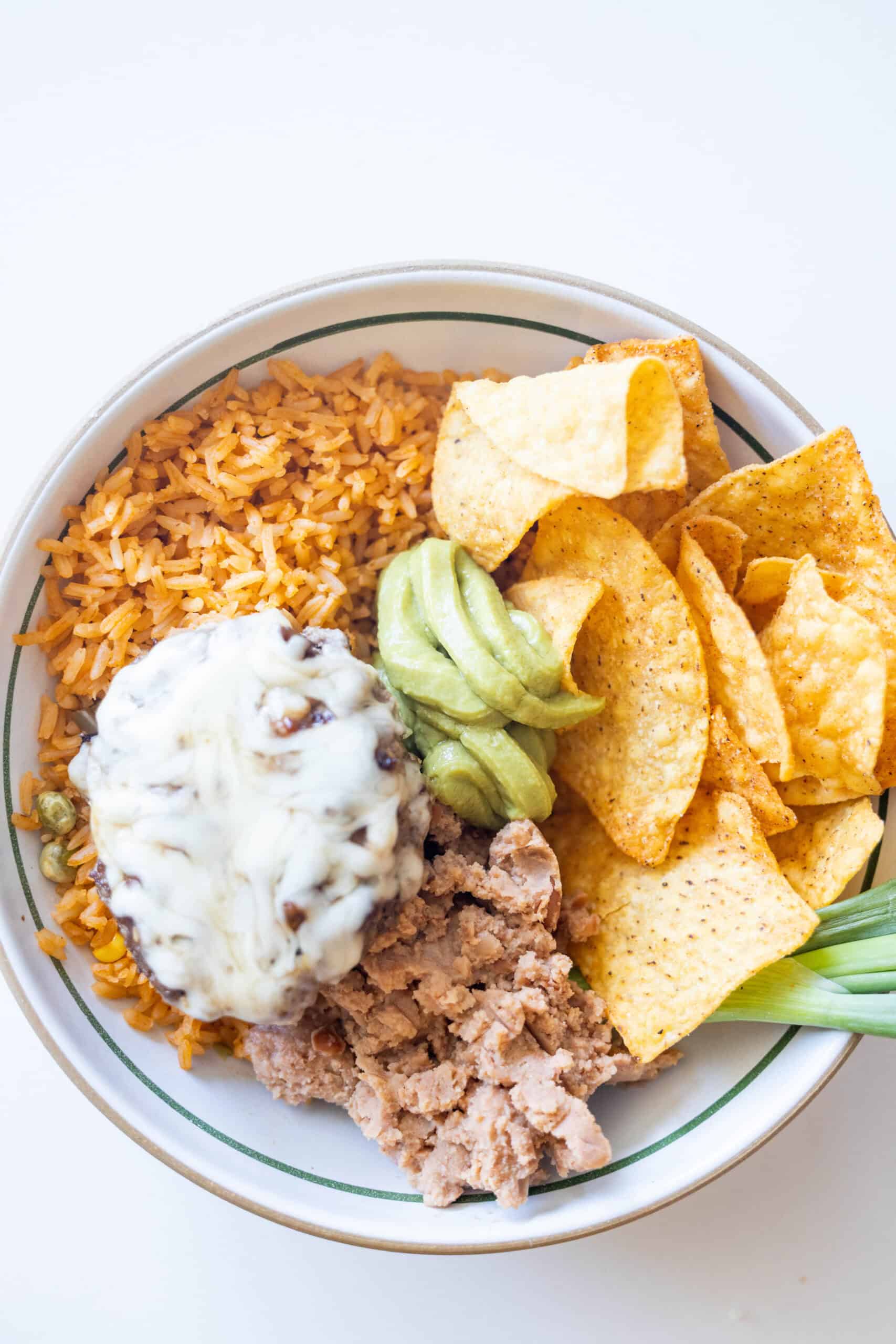 A bowl featuring Mexican rice, tortilla chips, refried beans, ground beef with melted cheese and green chile, topped with guacamole and garnished with a green onionโa flavorful twist on classic burger bowls.