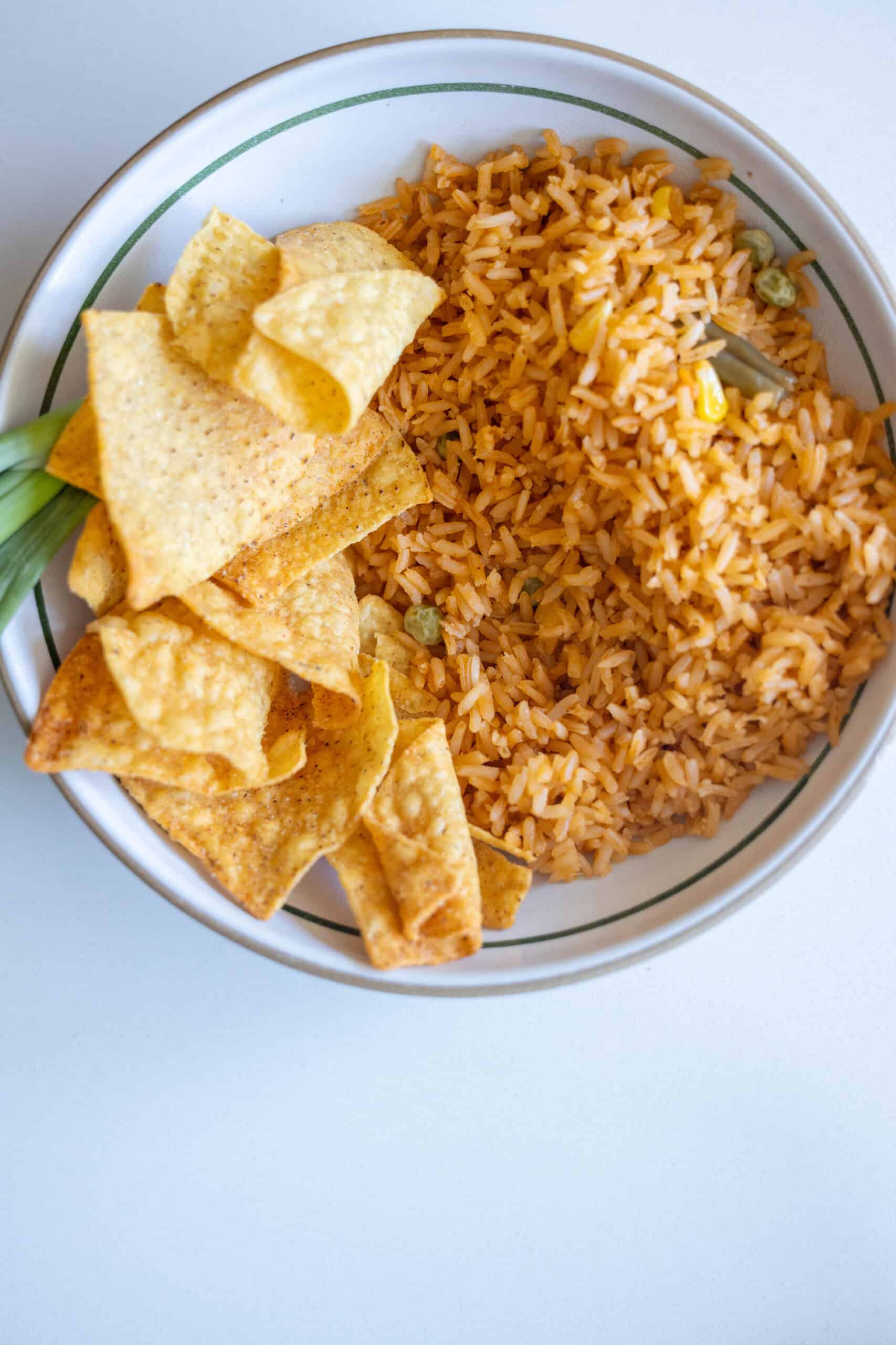 A bowl containing orange-colored rice with green peas and a hint of green chile, served alongside tortilla chips on a white surface.
