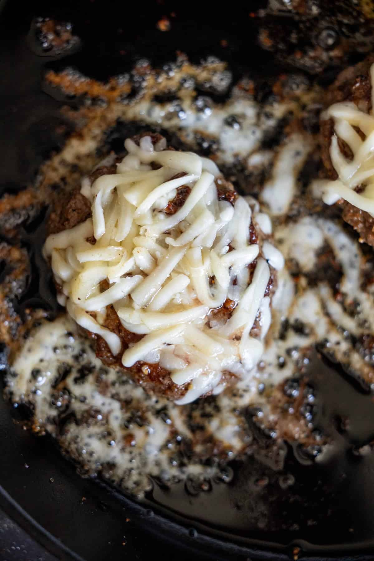 A cooked burger patty in a skillet topped with melted shredded cheese and zesty green chile, surrounded by grease and browned bits.
