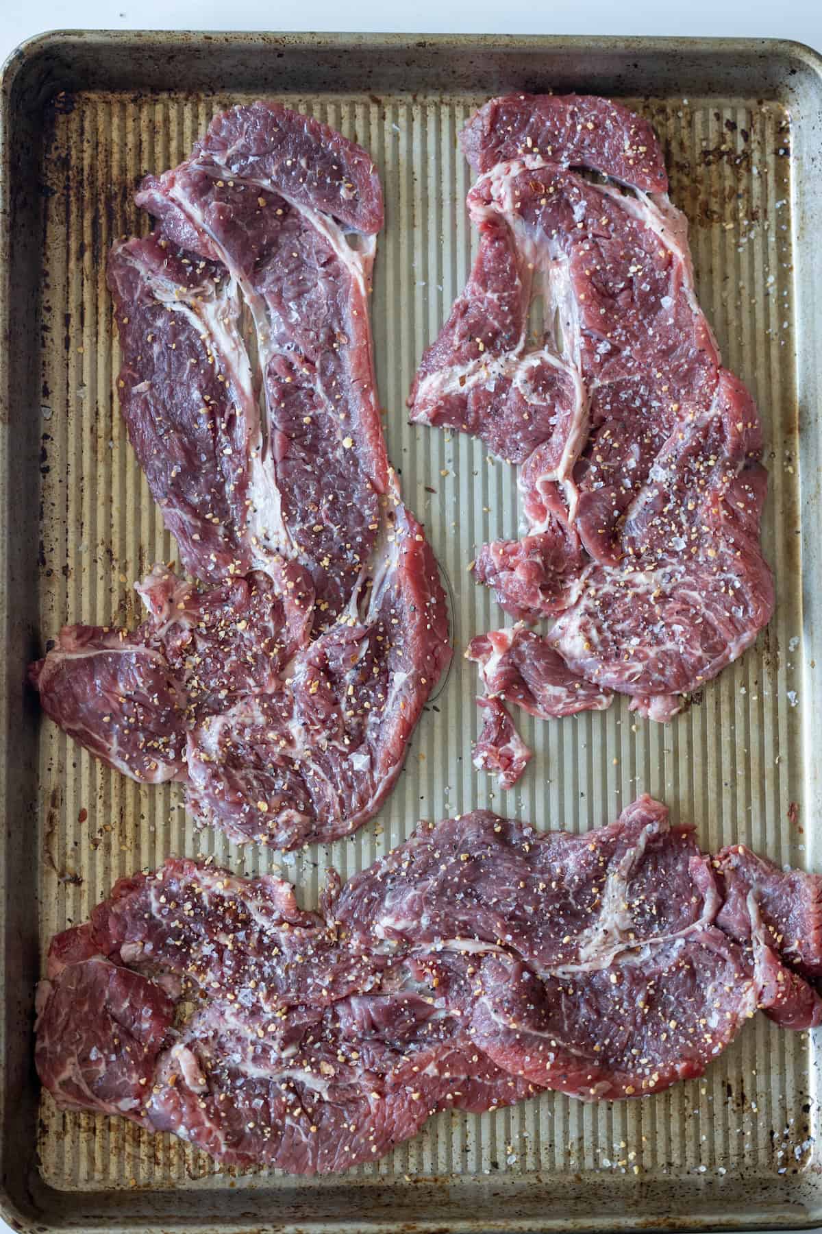 Three raw, marbled Armenian beef steaks seasoned with salt and pepper are arranged on a ridged metal baking sheet.