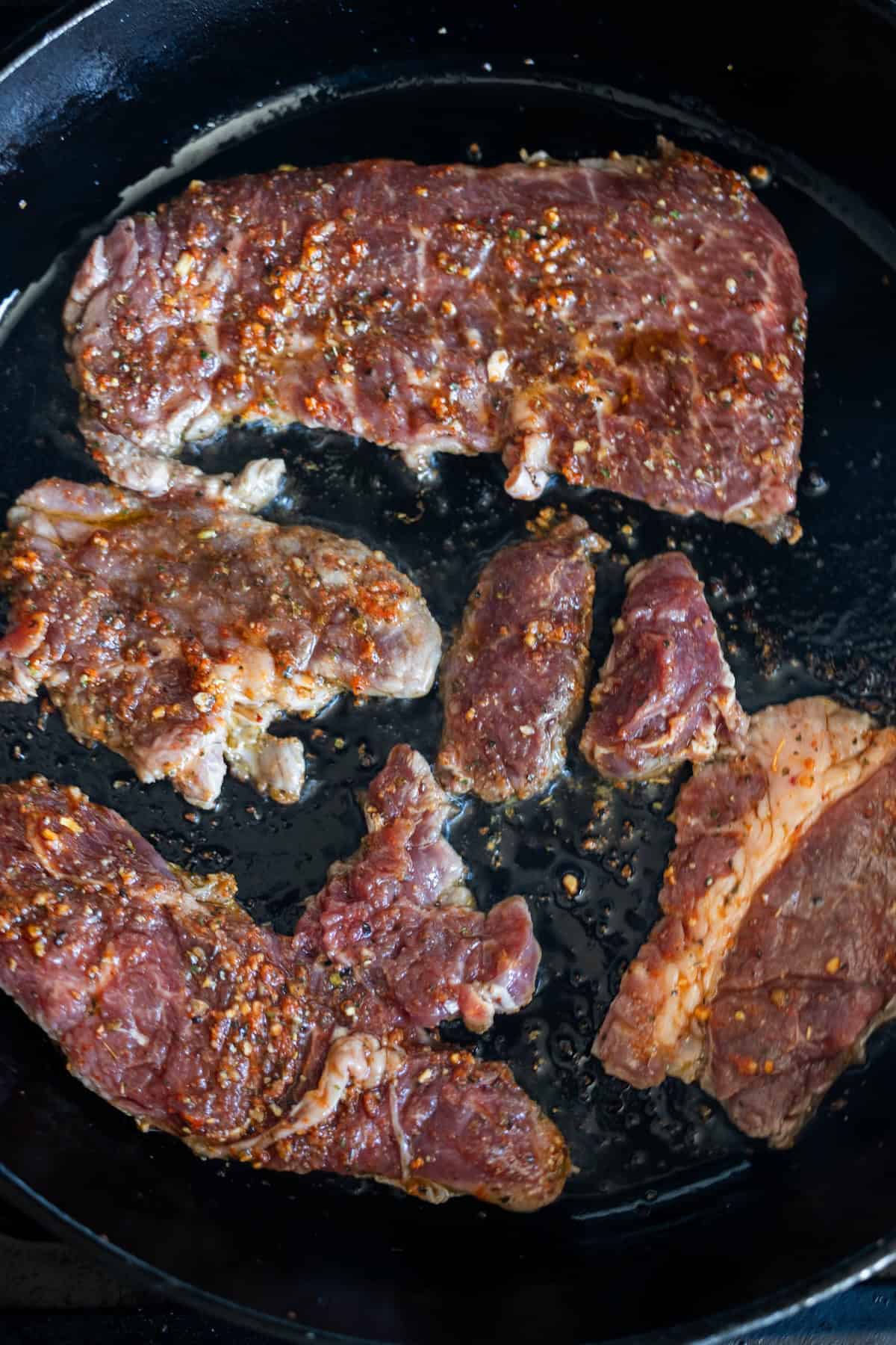 Slices of marinated Armenian beef are cooking in a black skillet, with visible spices and oil sizzling on the surface of the meat.