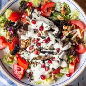 A bowl of Armenian salad featuring crisp lettuce, juicy tomato, crunchy walnuts, grilled Armenian beef, pomegranate seeds, and a creamy white tahini dressing.