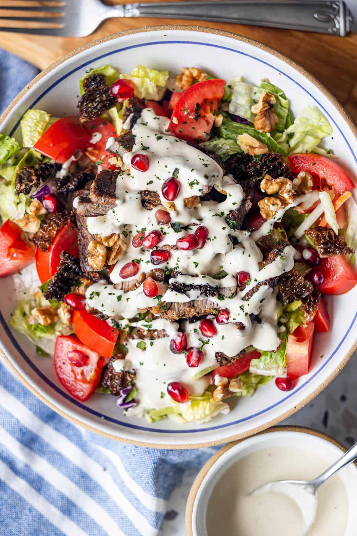 A bowl of Armenian salad with lettuce, tomatoes, walnuts, pomegranate seeds, tender grilled Armenian beef, and creamy dressing, served with utensils and a small bowl of extra tahini salad dressing nearby.