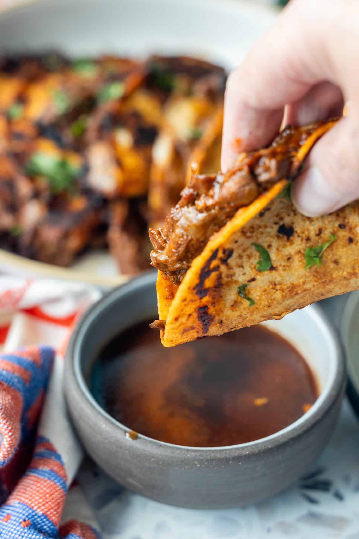 A hand dips a Birria Taco filled with tender meat into a bowl of rich consommé, with more Birria Tacos and a striped cloth visible in the background.