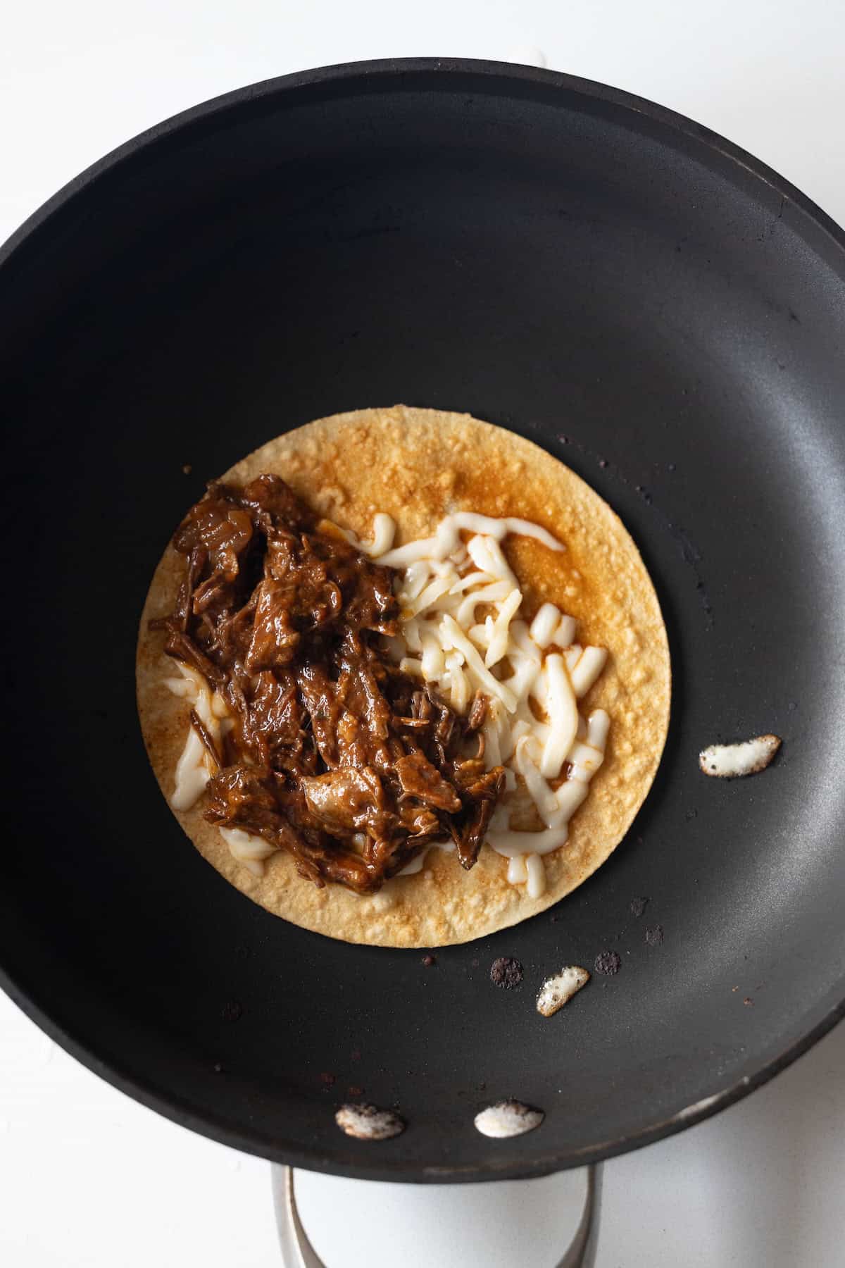 A corn tortilla in a black skillet is topped with shredded meat and grated cheese, capturing the mouthwatering essence of Birria Tacos, ready to be folded or cooked further.