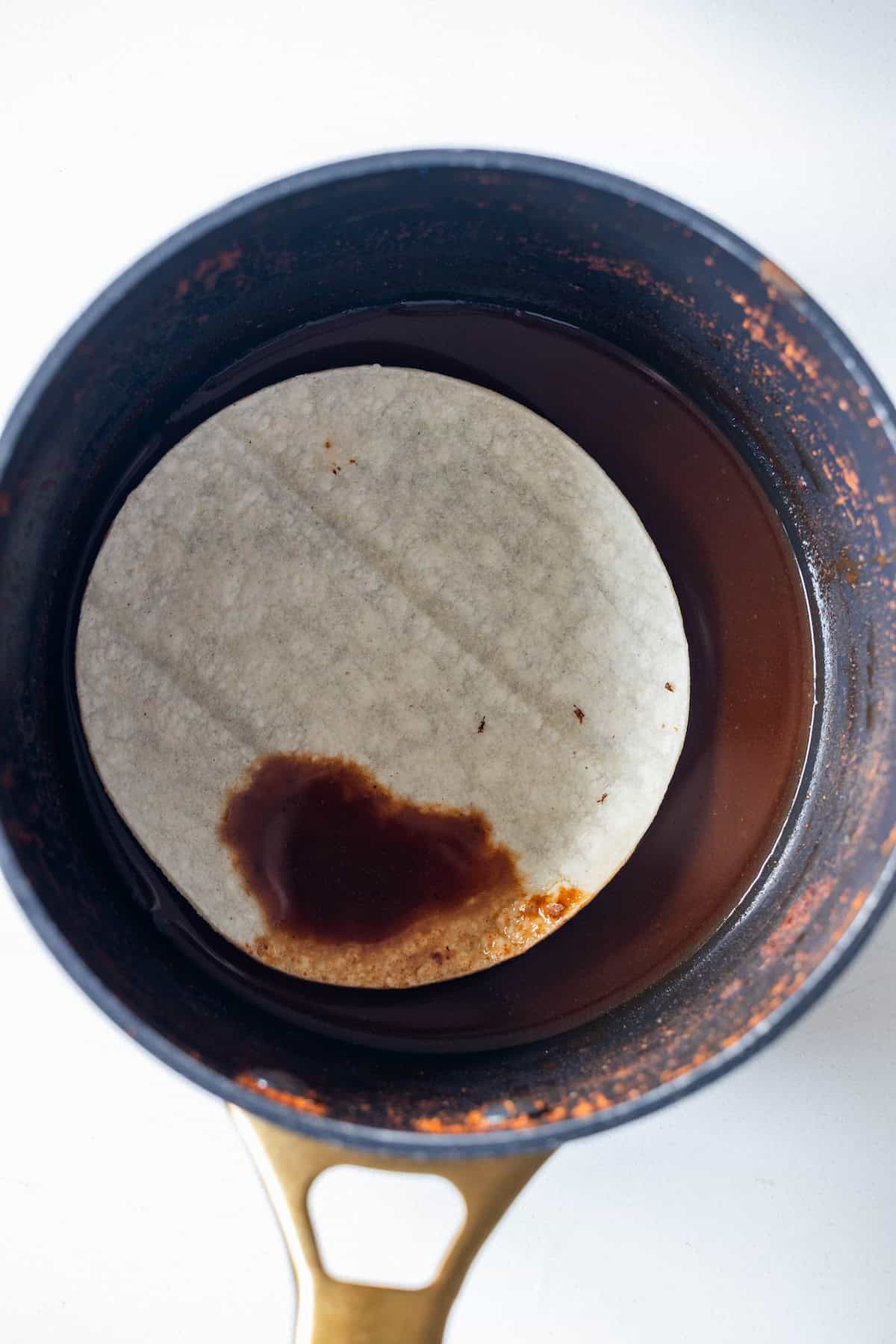 A corn tortilla, reminiscent of those used for Birria Tacos, is partially submerged in red sauce inside a small pot with a gold handle on a white surface.