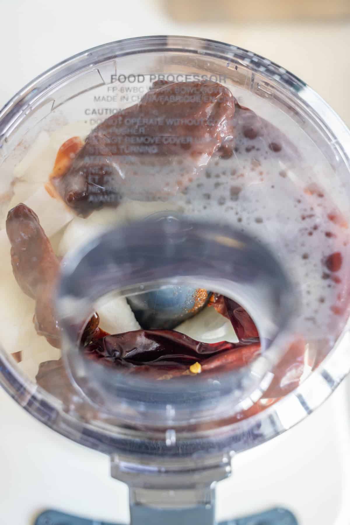 Top view of a food processor containing onions, dried red chilies, dates, and a foamy liquid—ingredients ready for blending to create the rich base often used in flavorful Birria Tacos.