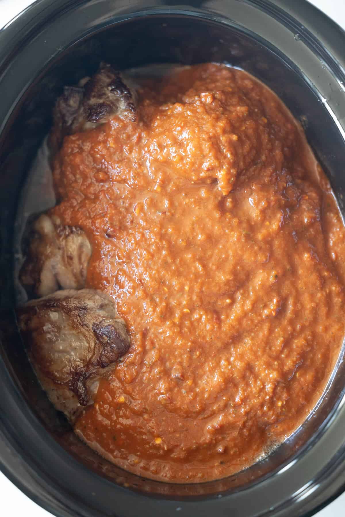 Slow cooker with browned meat partially covered in a thick red tomato-based sauce, perfect for making flavorful Birria Tacos.