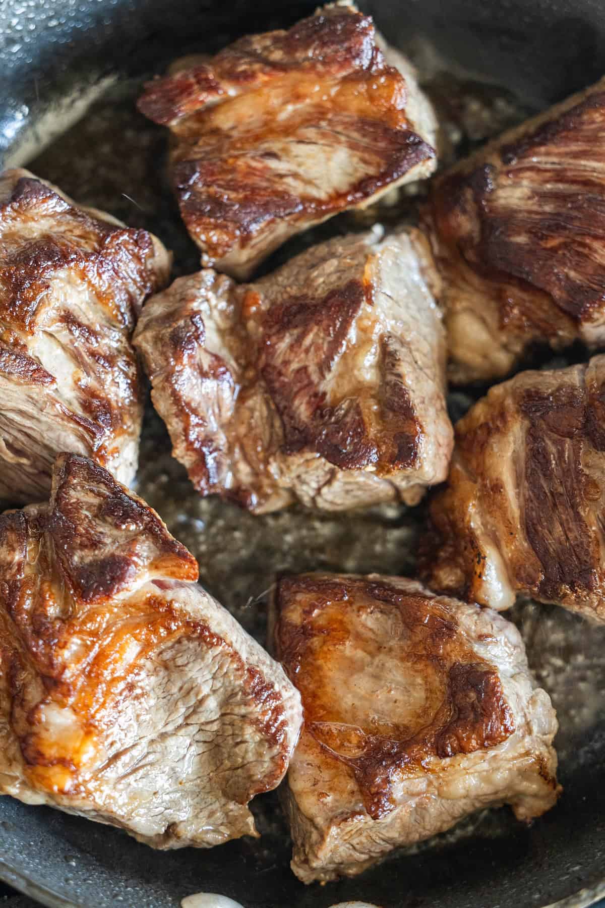 Several browned beef chunks are being seared in a black skillet, with visible caramelization on the surface of the meat—the perfect base for flavorful Birria Tacos.