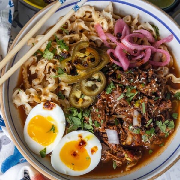 Bowl of Birria Ramen with shredded beef, soft-boiled egg halves, pickled onions, jalapeños, fresh herbs, and rich broth, served with chopsticks.