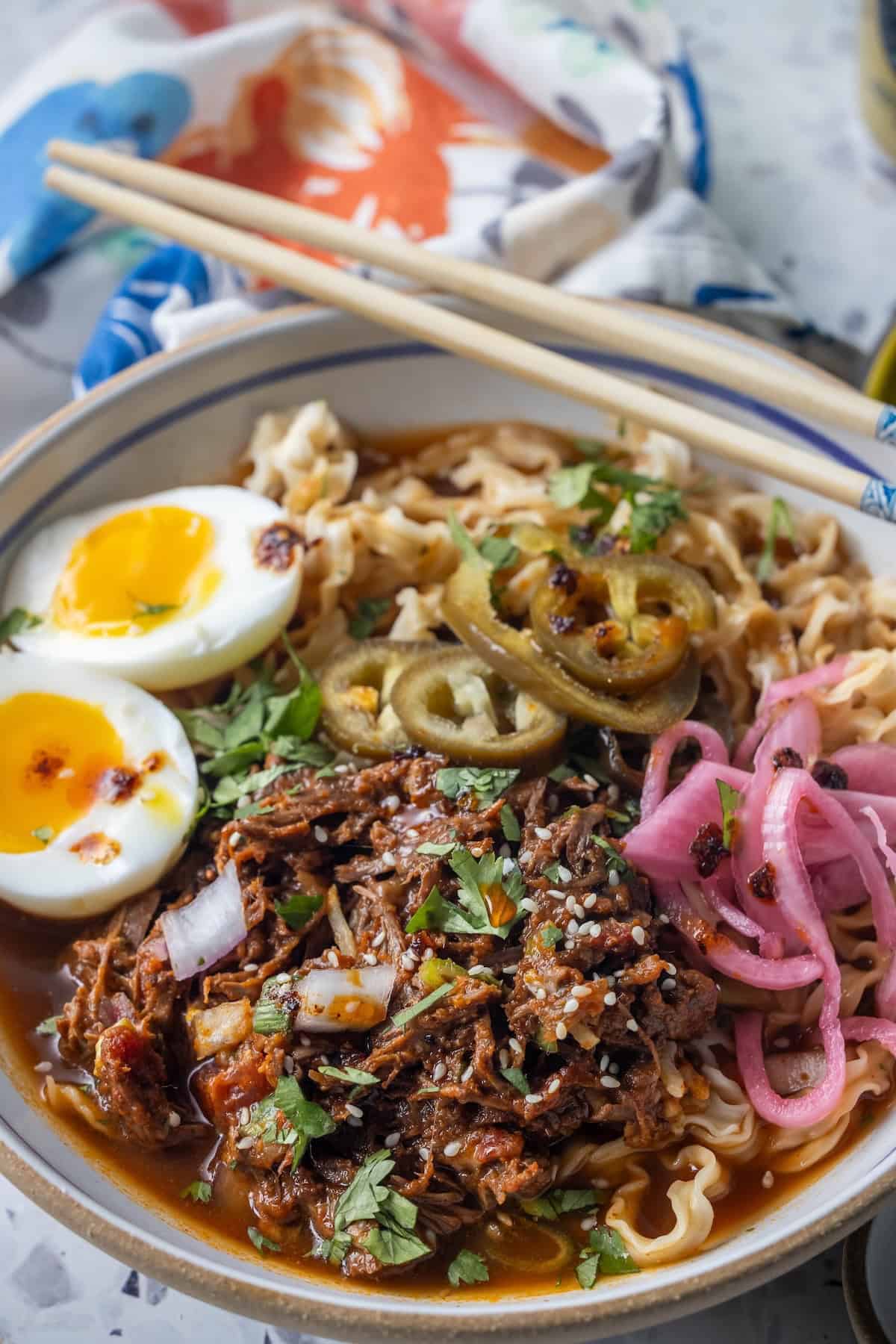 Bowl of Birria Ramen noodles topped with shredded meat, soft-boiled eggs, pickled onions, jalapeรฑos, chopped herbs, and sesame seeds, with chopsticks resting on the bowlโs rim.