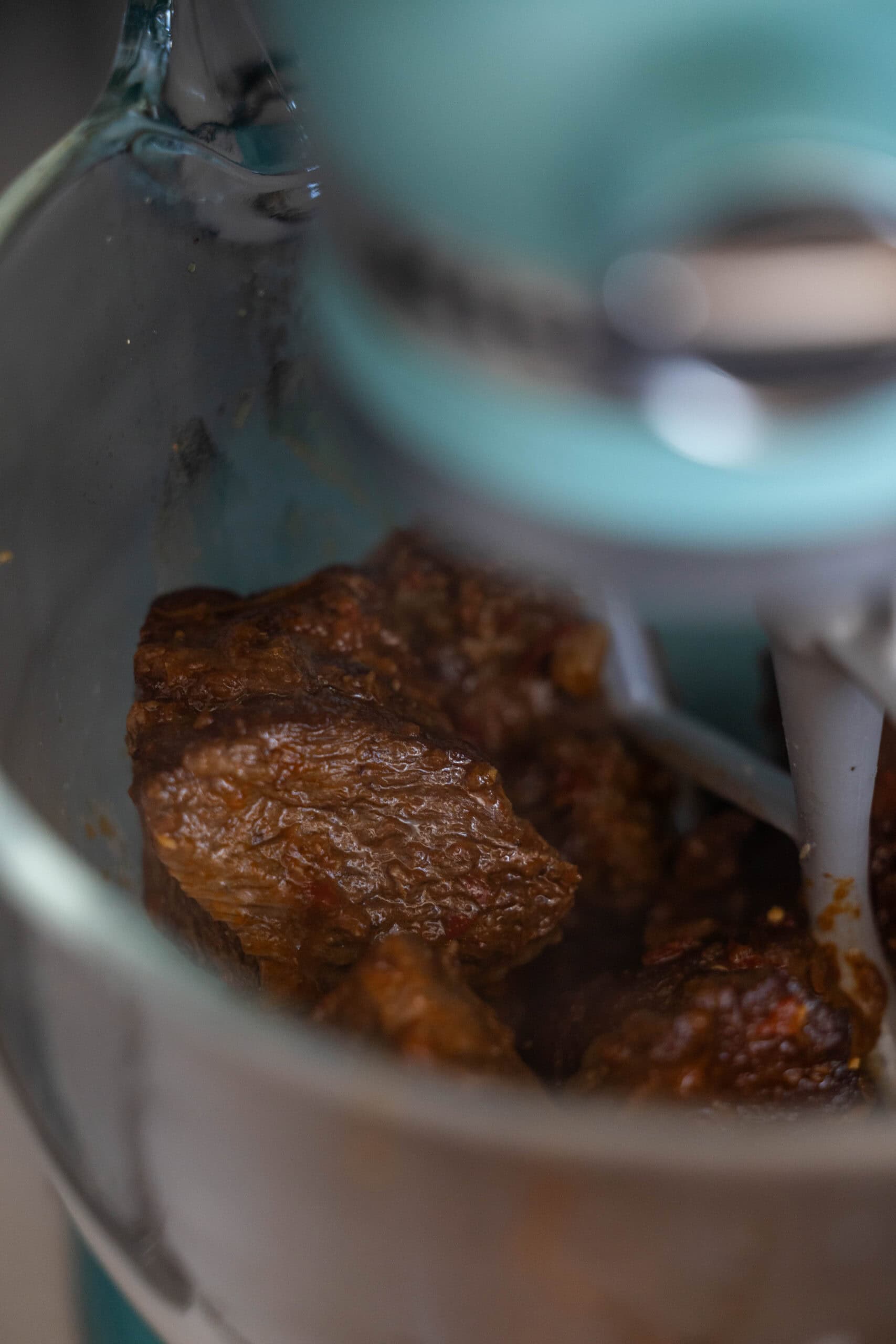 Close-up of chunks of cooked Birria being mixed in a stand mixer with a metal paddle attachment inside a glass mixing bowl.