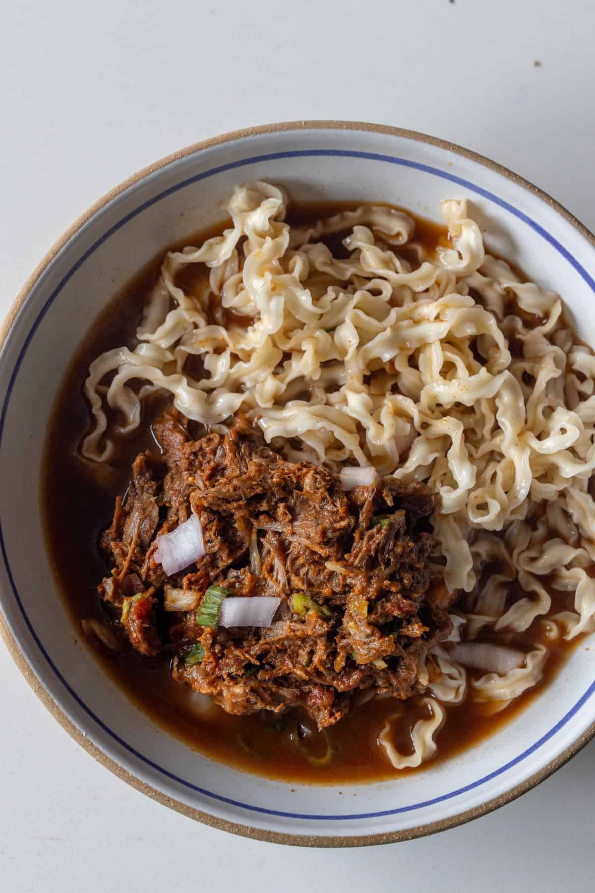 A bowl of wavy ramen noodles in broth topped with flavorful birria-style shredded meat and chopped onions, photographed from above on a white background.