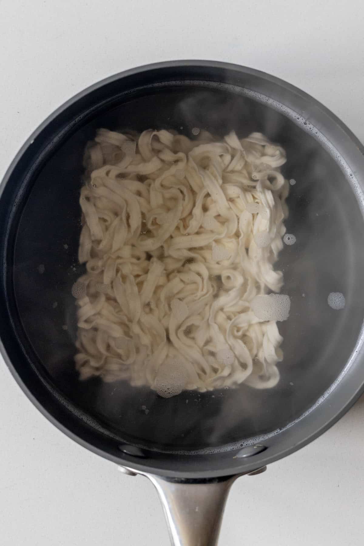 A black pot of boiling water with uncooked, flat, white noodles being cooked, perfect as a base for a flavorful Birria Ramen.