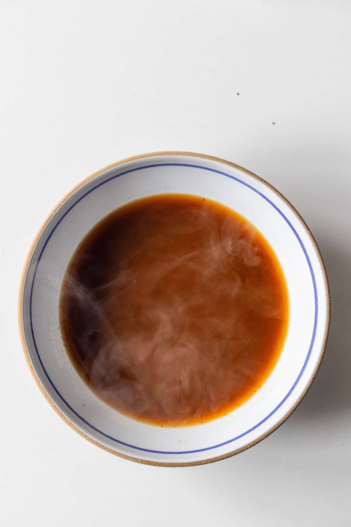 A bowl of hot, steaming Birria Ramen sits on a white surface, its rich brown broth inviting you to savor the fusion of traditional Birria flavors with comforting ramen noodles.
