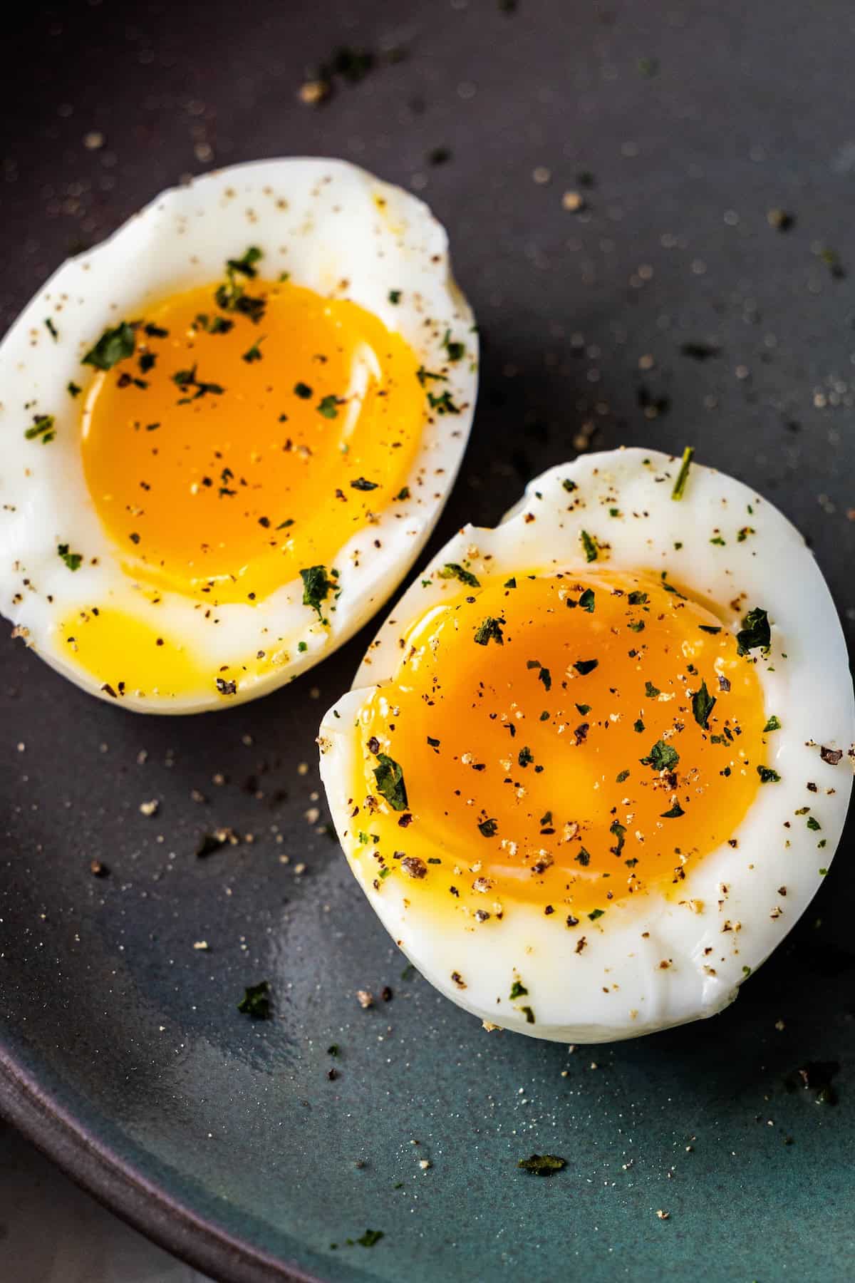 Two soft-boiled egg halves with runny yolks, sprinkled with black pepper and herbs, on a dark plateโperfect as a savory topping for your favorite bowl of ramen.