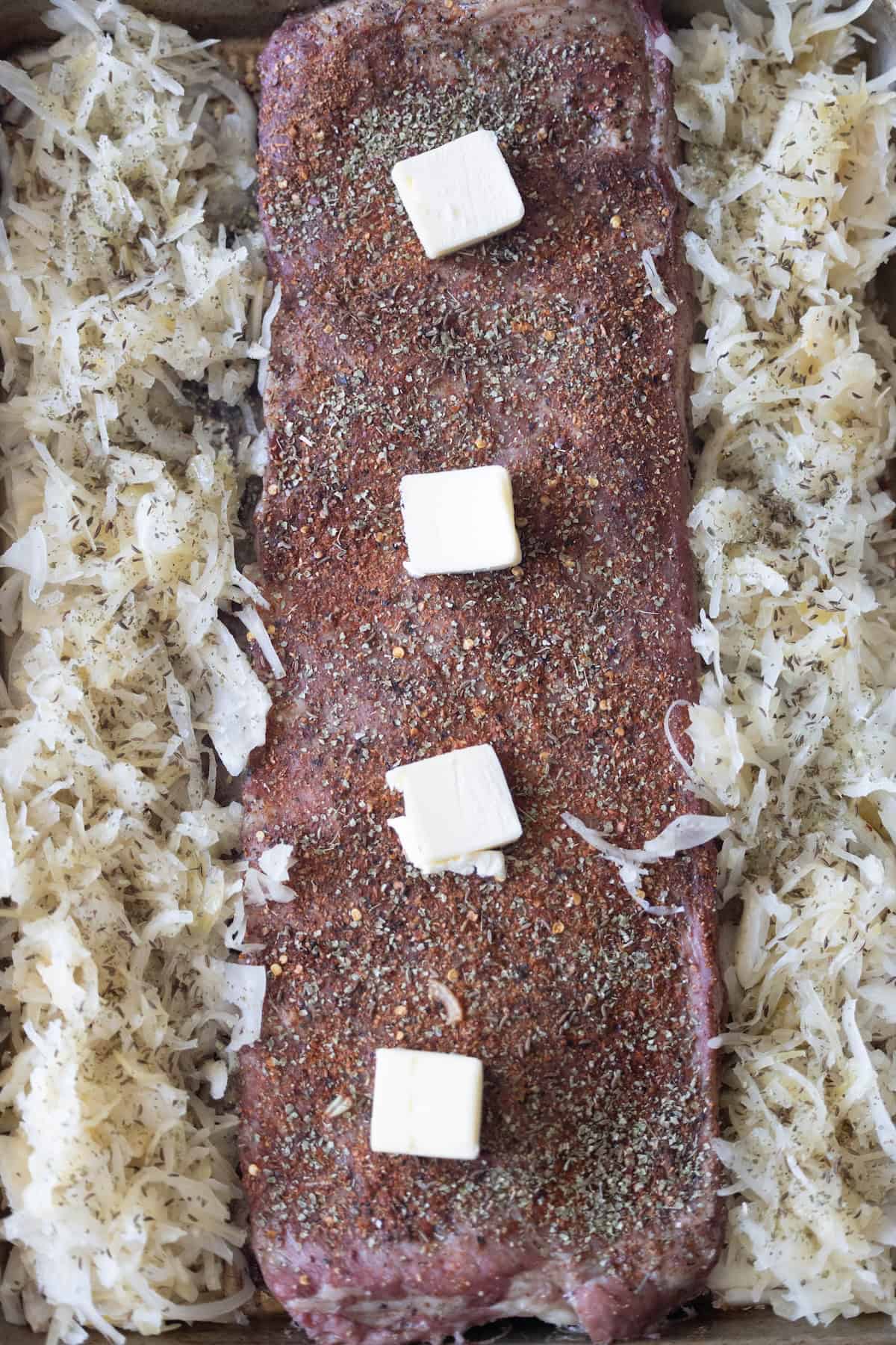 Uncooked seasoned pork loin topped with four butter cubes, surrounded by shredded sauerkraut in a baking dish, ready for the ovenโan inspired take on a classic roasted ribs recipe.