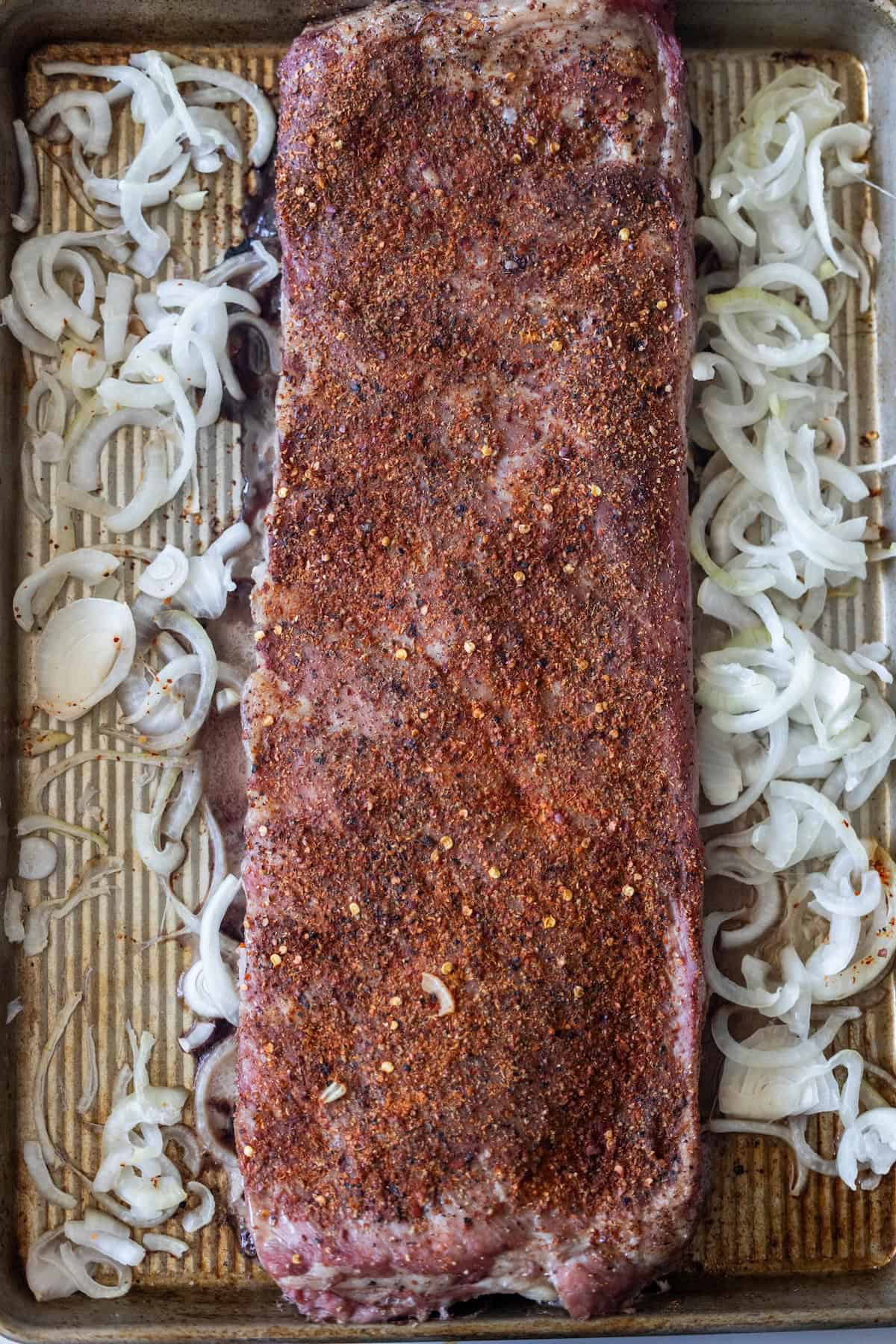 A rack of seasoned pork ribs on a baking tray, surrounded by sliced raw onions, ready to become the centerpiece of your favorite roasted ribs recipe. For an extra layer of flavor, try pairing these tender ribs with tangy sauerkraut.