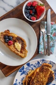 A plate with rich Challah French Toast topped with butter and berries, a bowl of mixed berries, utensils on a napkin, and another plate with more French Toast on a wooden board.