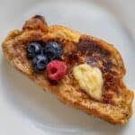 A slice of rich Challah French Toast is topped with a pat of butter, three blueberries, and one raspberry on a white plate.