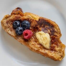 A slice of rich Challah French Toast is topped with a pat of butter, three blueberries, and one raspberry on a white plate.