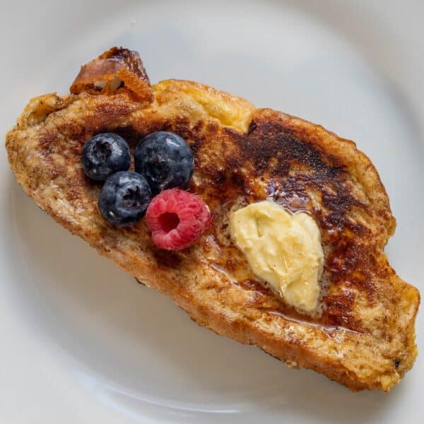 A slice of rich Challah French Toast is topped with a pat of butter, three blueberries, and one raspberry on a white plate.