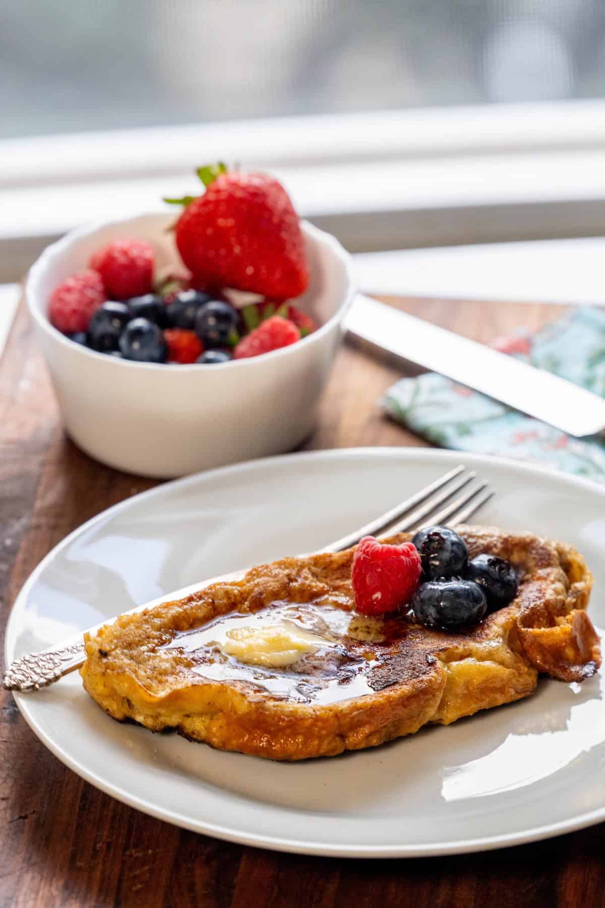 A slice of cinnamon sugar French toast made with rich challah bread sits on a white plate, topped with butter, blueberries, and a raspberry, alongside a fork; a bowl of mixed berries is in the background on a wooden table.
