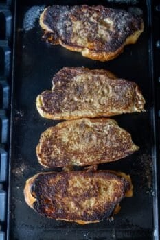 Four slices of French toast are cooking on a griddle, made from thick-cut Challah and topped with cinnamon sugar, with visible browning and crispy edges on each piece.