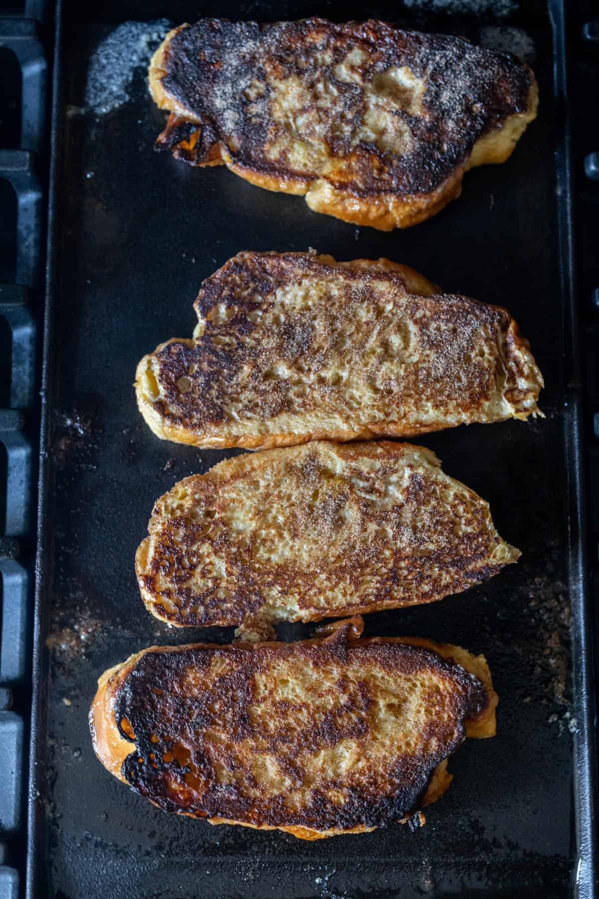 Four slices of French toast are cooking on a griddle, made from thick-cut Challah and topped with cinnamon sugar, with visible browning and crispy edges on each piece.