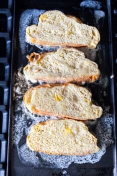 Four slices of bread coated in egg mixture are cooking on a buttered griddle, making classic French toast with rich Challah bread and a hint of cinnamon sugar.