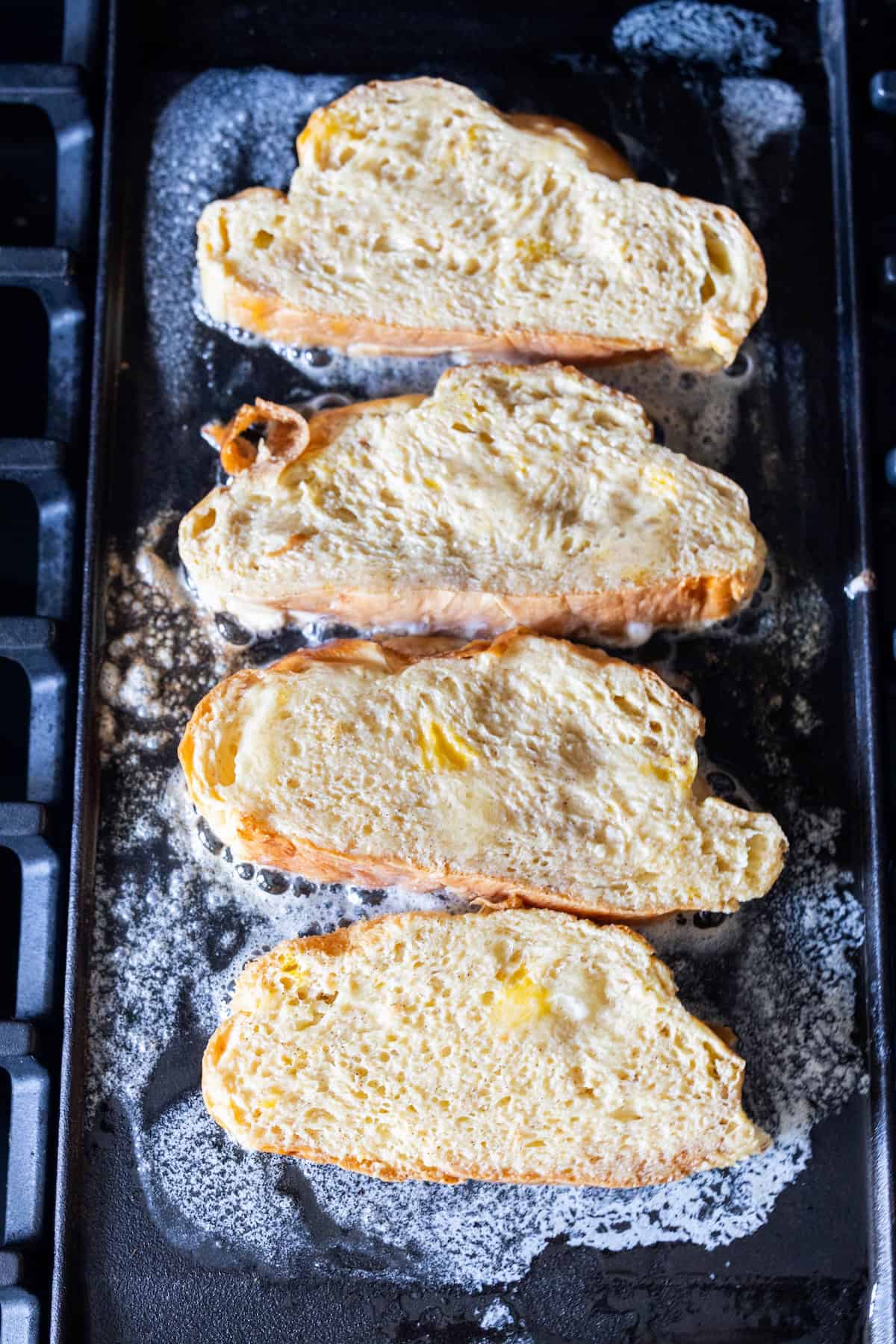 Four slices of bread coated in egg mixture are cooking on a buttered griddle, making classic French toast with rich Challah bread and a hint of cinnamon sugar.