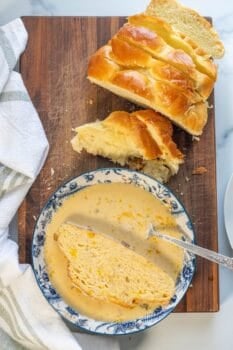 A slice of braided bread in a bowl of soup with a spoon, next to more sliced bread on a wooden cutting board with a white towel nearby&mdash;perfect for transforming into leftover challah French toast the next morning.
