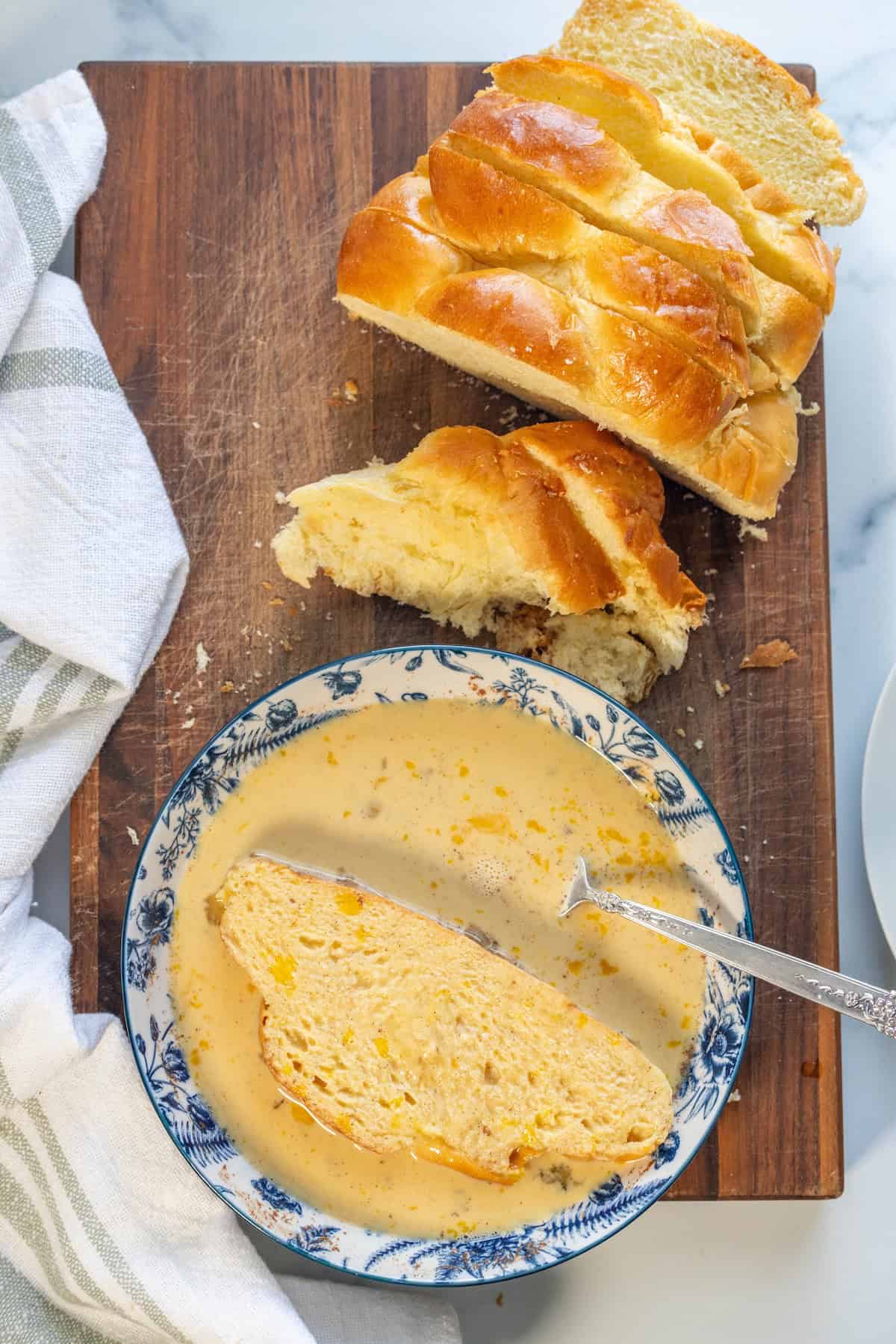 A slice of braided bread in a bowl of soup with a spoon, next to more sliced bread on a wooden cutting board with a white towel nearby&mdash;perfect for transforming into leftover challah French toast the next morning.