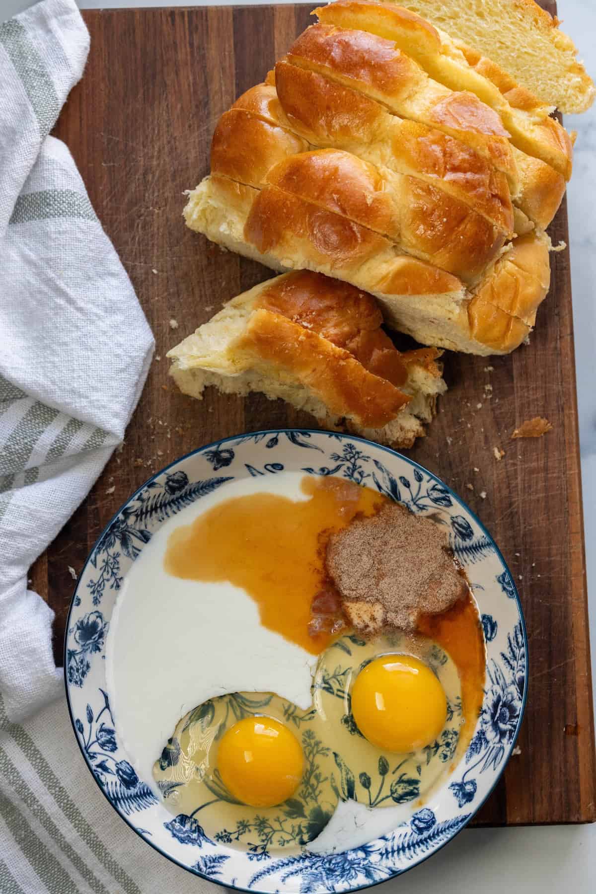 Sliced braided challah bread on a wooden board next to a bowl with two eggs, milk, sugar, and vanilla extract&mdash;perfect ingredients for making French Toast. A white-striped towel is on the side.