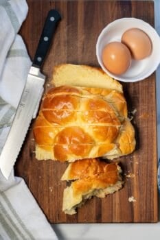 A loaf of braided challah bread is sliced on a wooden cutting board with a serrated knife, next to a white bowl holding two brown eggs and a striped kitchen towel&mdash;perfect for making cinnamon sugar French toast.
