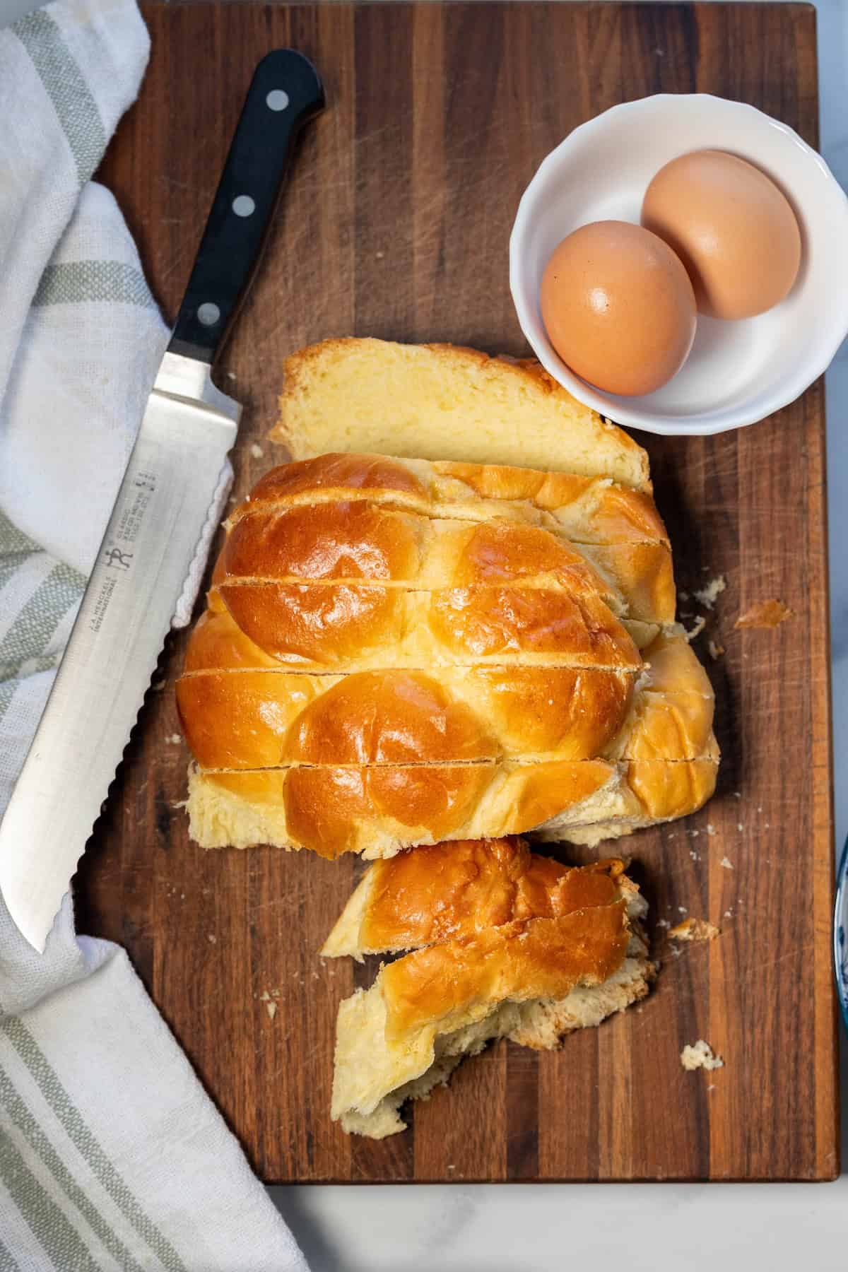 A loaf of braided challah bread is sliced on a wooden cutting board with a serrated knife, next to a white bowl holding two brown eggs and a striped kitchen towel&mdash;perfect for making cinnamon sugar French toast.