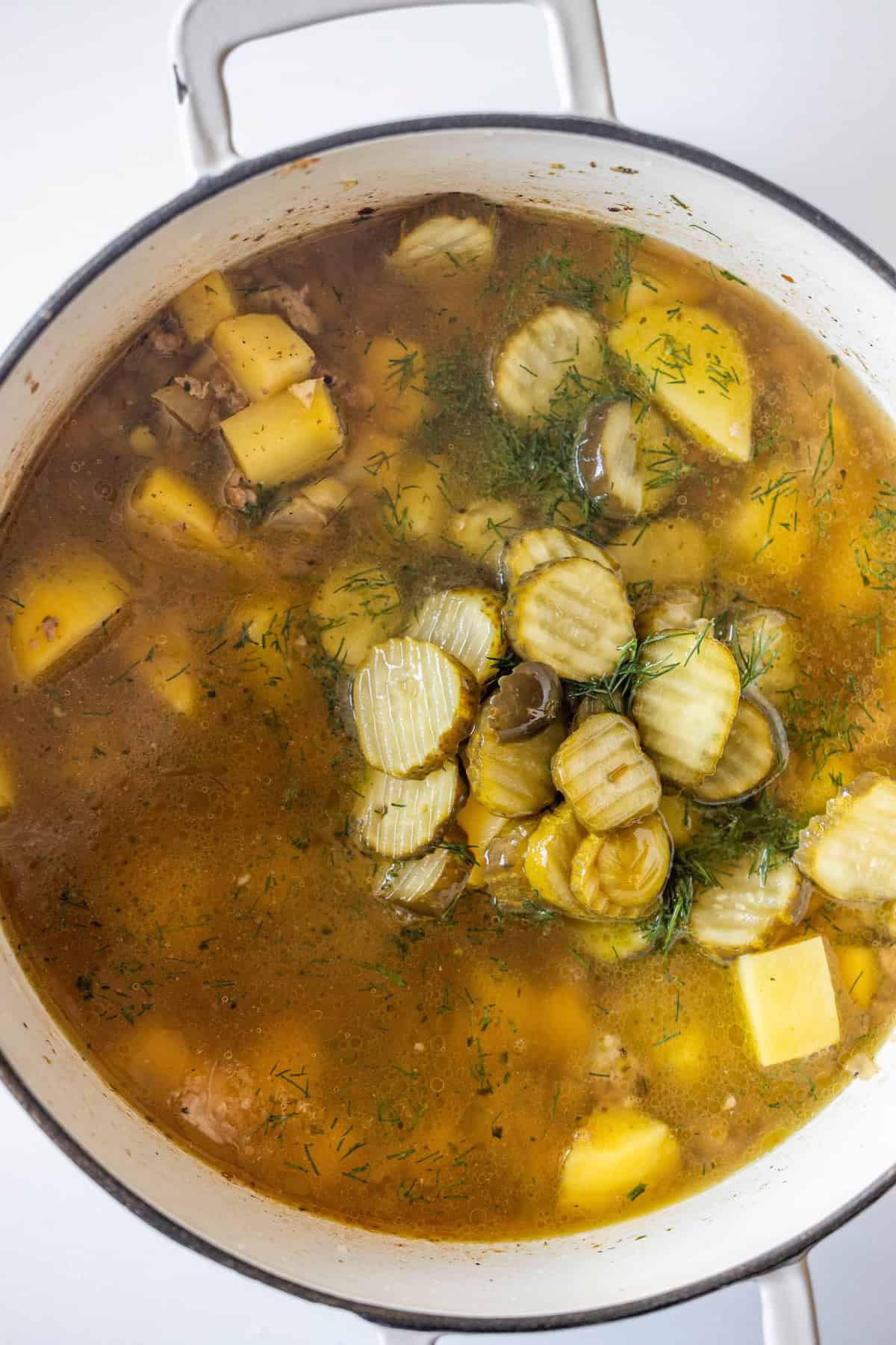 A pot of soup with chunks of potato, sliced pickles, savory sausage, herbs, and broth, viewed from above.