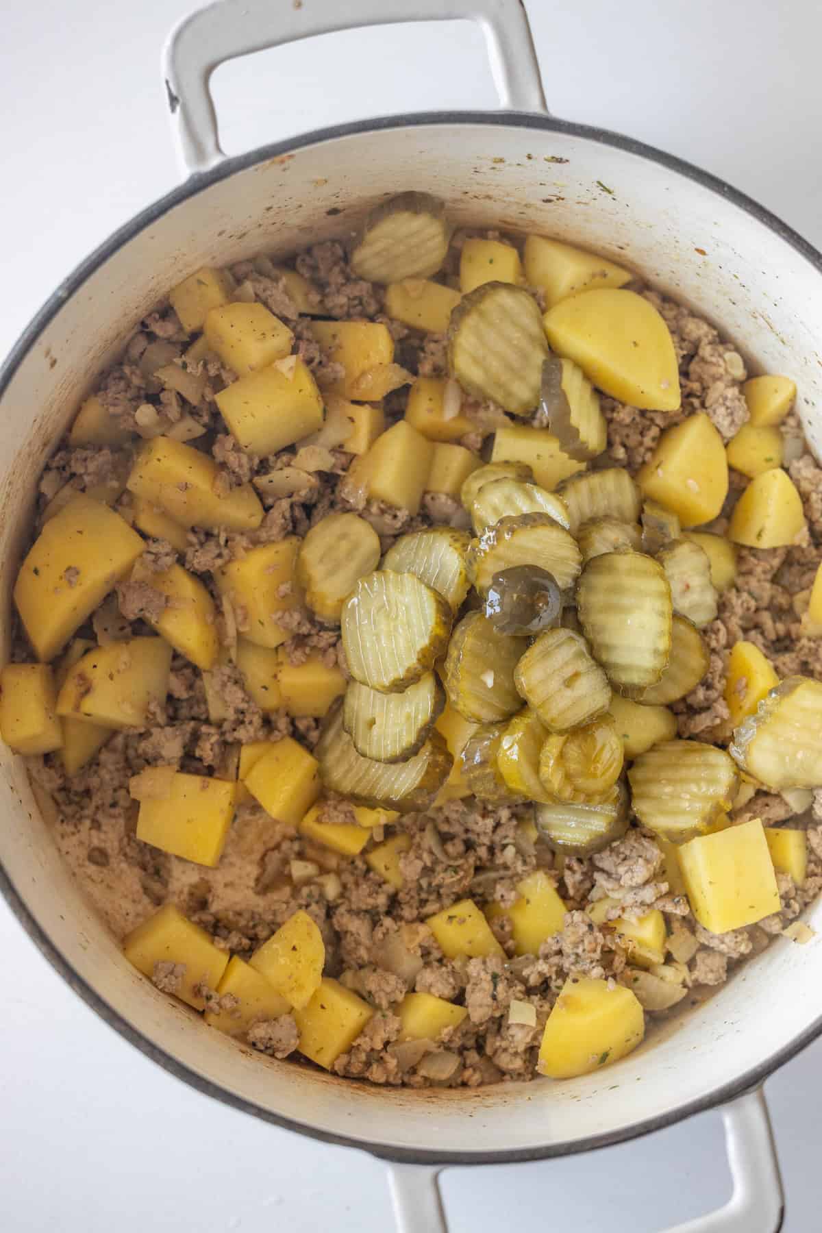 A pot containing cooked ground meat, diced potatoes, sliced pickles, and pieces of sausage, all mixed together on a white surface.